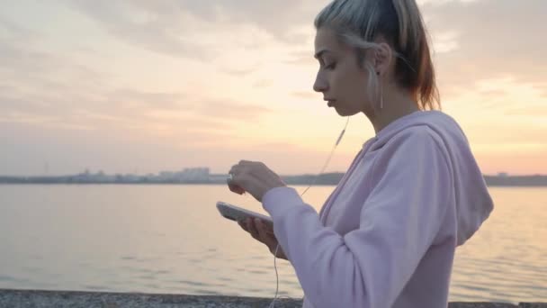 Jong meisje in hoofdtelefoon loopt op een achtergrond van zonsondergang en water, slow motion — Stockvideo