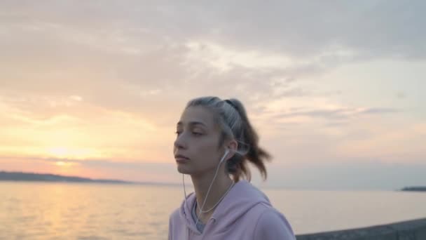 Mladá dívka ve sluchátkách běží na pozadí západu slunce a vody, zpomalení — Stock video