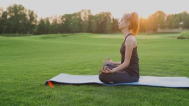 Genç kız lotus pozisyonunda yeşil çimlere oturur, gün batımında yoga dersi..