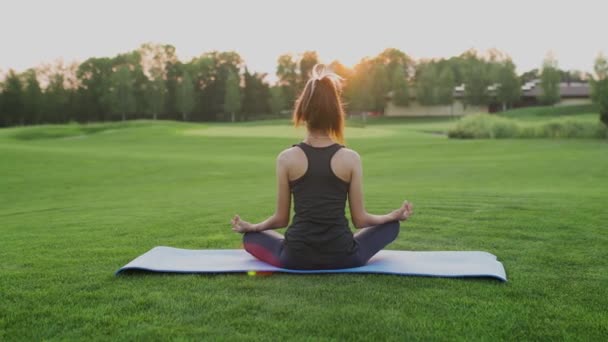 Ung flicka sitter på grönt gräs i lotusposition, yoga klass vid solnedgången tid. — Stockvideo