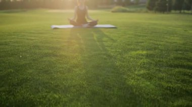 Genç kız lotus pozisyonunda yeşil çimlere oturur, gün batımında yoga dersi..