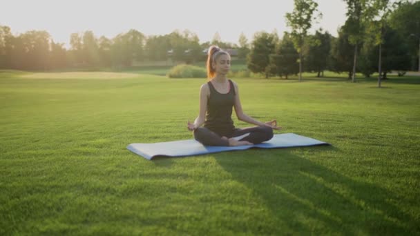 Jong meisje zit op groen gras in lotus positie, yoga klasse bij zonsondergang tijd. — Stockvideo