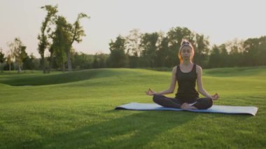 Genç kız lotus pozisyonunda yeşil çimlere oturur, gün batımında yoga dersi..