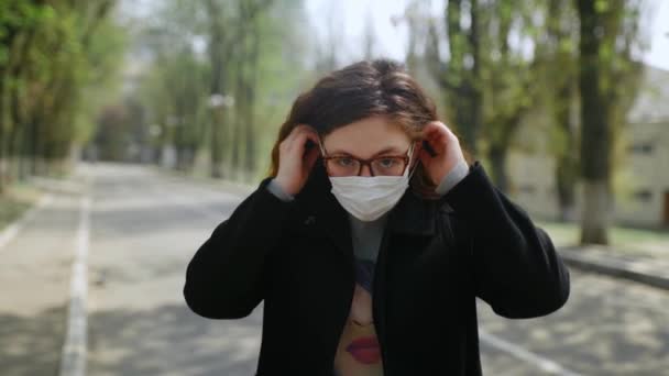 A young red-haired girl stands on the street, puts on a medical mask and looks at the camera. — Stock Video