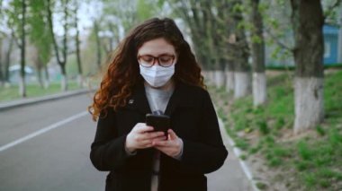 Tıbbi maskeli genç bir kız parkta akıllı telefon kullanıyor. 4k