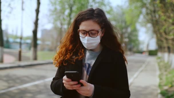 Chica joven con una máscara médica utiliza un teléfono inteligente en el parque. 4k — Vídeo de stock