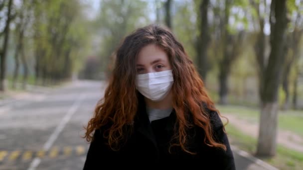 Молодая девушка в медицинской маске стоит на улице в парке. Концепция COVID-19 — стоковое видео