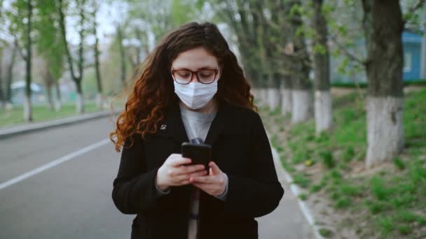 Ung flicka i medicinsk mask använder en smartphone i parken. 4k — Stockvideo