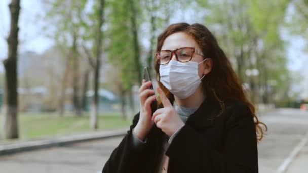Rapariga em uma máscara médica usa um smartphone no parque. 4k — Vídeo de Stock
