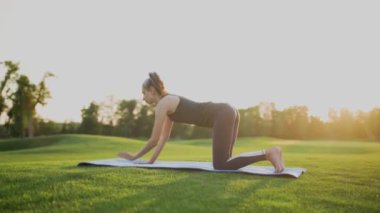 Yeşil çimenlerin üzerinde bir parkta genç bir kız bir tahtada duruyor, gün batımında spor yapıyor..