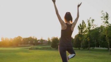 Genç bir kız gün batımında yeşil çimlerin üzerinde durmuş yoga yapıyor.