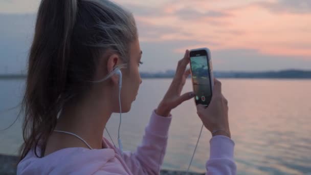 女の子は水と日没の背景にウォーターフロントの電話を使用します。. — ストック動画