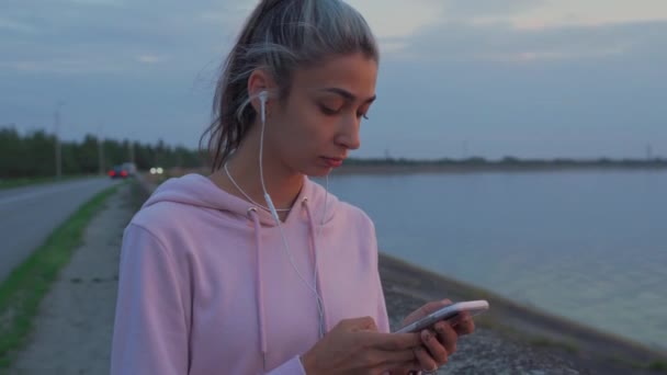 Chica caminando por el paseo marítimo escribe un mensaje en un teléfono inteligente . — Vídeo de stock