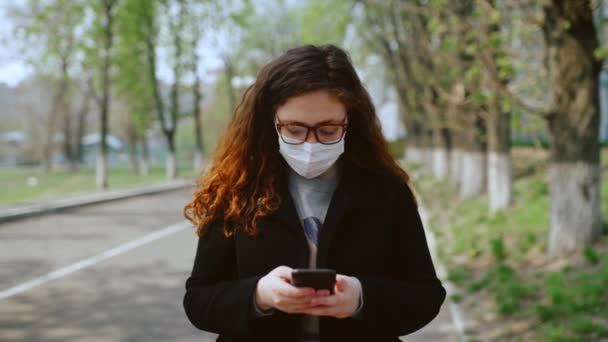 Chica joven con una máscara médica utiliza un teléfono inteligente en el parque. 4k — Vídeos de Stock