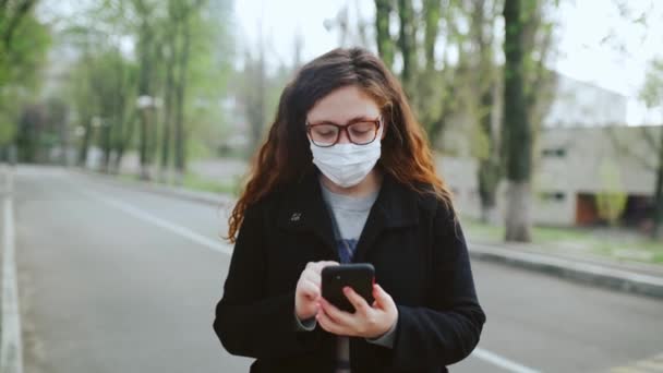 Chica joven con una máscara médica utiliza un teléfono inteligente en el parque. 4k — Vídeos de Stock