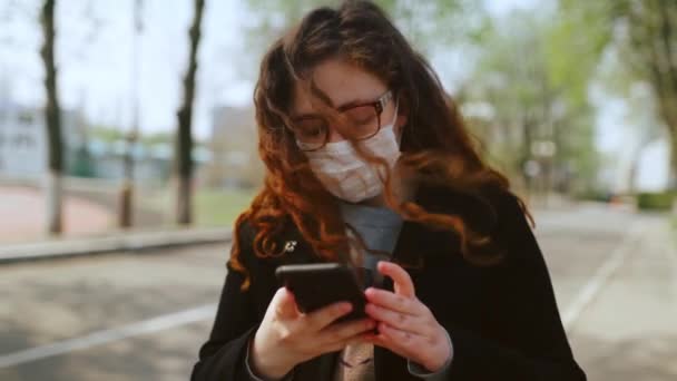 Chica joven con una máscara médica utiliza un teléfono inteligente en el parque. 4k — Vídeo de stock