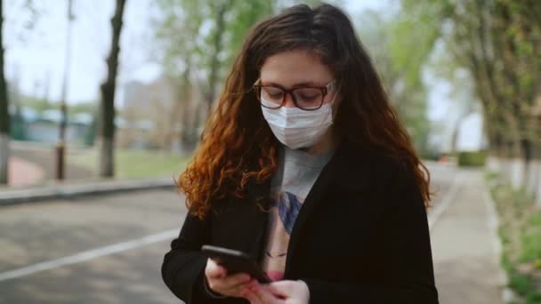 Chica joven con una máscara médica utiliza un teléfono inteligente en el parque. 4k — Vídeos de Stock