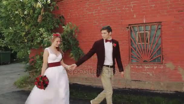 Braut und Bräutigam gehen an einer ziegelroten Wand entlang — Stockvideo
