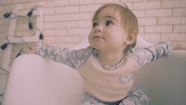 Alegre niña morena pequeña sentada en silla blanca con sonrisa cámara lenta — Vídeo de stock