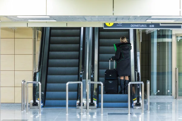 The alone woman on the escalator or moving staircase with inscription departure in English and Chinese in the international airport or railway station from the back moving upstairs with luggage