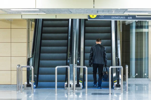The alone man on the departure escalator or moving staircase in the international airport or railway station from the back moving upstairs with luggage