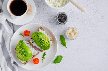 Avokado sandviçi ve koyu çavdar ekmeği taze avokado, krem peynir ve tohumlar beyaz tabakta ve açık arkaplanda bir fincan kahve. Yatay, üst görünüm, boşluğu kopyala.