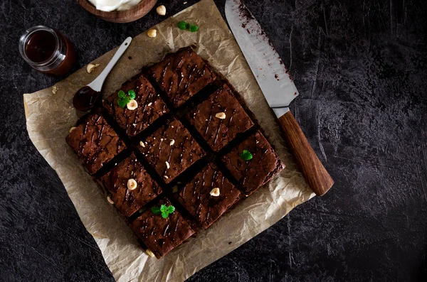 Домашній шоколадний десерт брауні з лісовими горіхами, шоколадною глазур'ю та збитими вершками на папері для випічки на темному фоні. Вид зверху. Горизонтальна орієнтація. Копіювати простір . — стокове фото