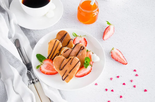 Herzförmige Pfannkuchen mit Schokoladensauce und frischen Erdbeeren auf einem weißen Teller vor hellem Hintergrund mit einer Tasse Kaffee. Frühstück zum Valentinstag. horizontale Orientierung. Kopierraum. — Stockfoto