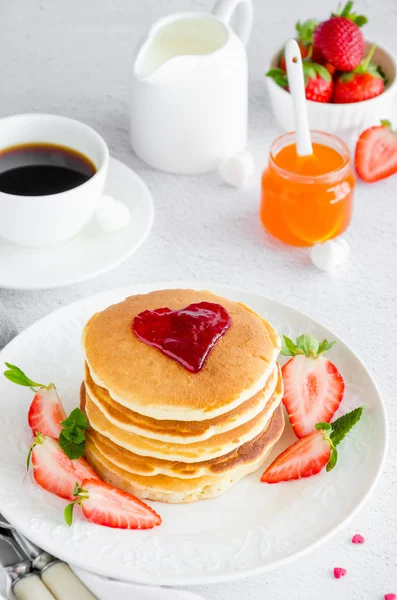 En bunt pannkakor med ett hjärta av sylt ovanpå med färska jordgubbar och mynta på en vit tallrik på en lätt bakgrund. Frukost till alla hjärtans dag. Vertikal orientering. Kopiera utrymme — Stockfoto