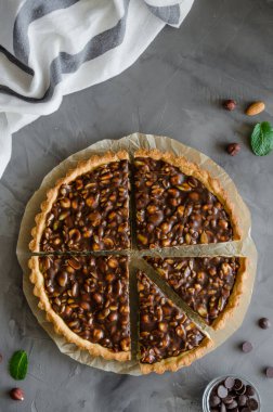 Çikolatalı karamelli tart, fındık, fıstık, badem ve tohum karışımı koyu beton bir arka planda. Dikey oryantasyon. Üst görünüm