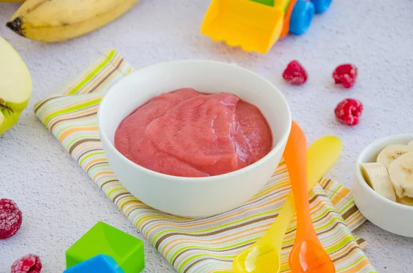 Baby food. Homemade apple puree or sauce with banana and raspberries in a bowl with a spoon on a light background. Healthy food. Horizontal orientation