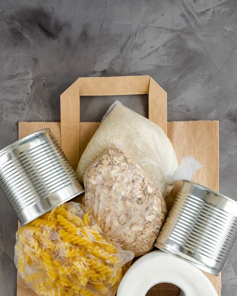 Donation food. Paper bag with canned food, pasta, oatmeal, rice and toilet paper on a dark concrete background. Food delivery. Vertical, top view, copy space