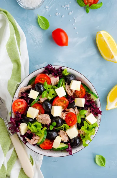Salad with tuna, fresh vegetables, olives and feta cheese in a bowl on a light blue concrete background. Healthy food. Vertical orientation, top view