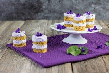 Menekşe çiçekli küçük pastalar, eski ahşap masaya serilmiş maskara krema.