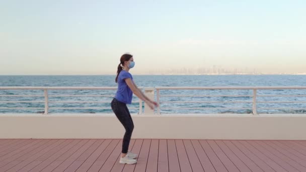 Chica en máscara médica hace sentadillas frente al mar — Vídeos de Stock