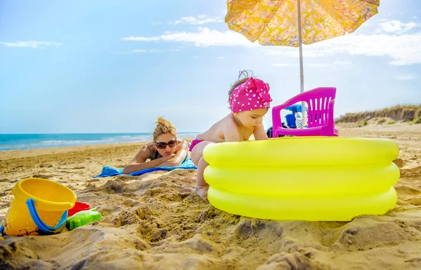 小女孩玩黄色充气泳池妈妈在沙滩巾上检查她的日光浴 — 图库照片