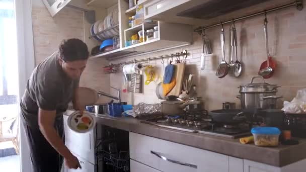 Мужчина выполняет рутинную работу на кухне квартиры, загружая посудомоечную машину и готовя еду для дочери на газовой плите — стоковое видео