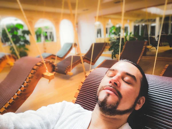 Man in spa relax area on suspended deck chair alone in silence with eyes closed and bathrobe — Stock Photo, Image