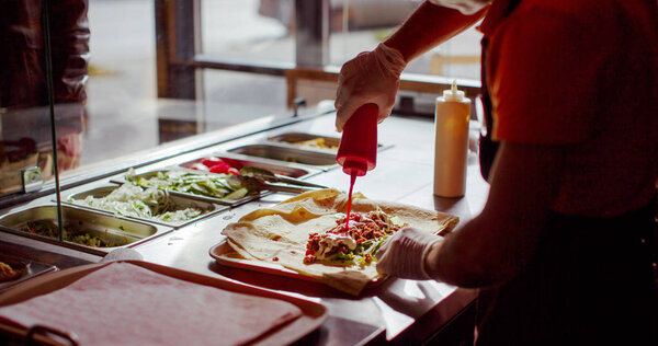 Crop person in gloves pouring ketchup in burrito while cooking in cafe.