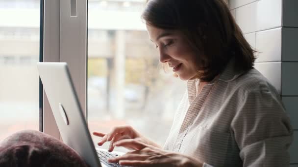 Pencere üzerinde oturan bir dizüstü bilgisayarda yazarak kız — Stok video