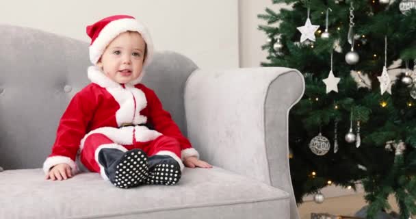 Baby i santa kostym sit soffan med julgran — Stockvideo