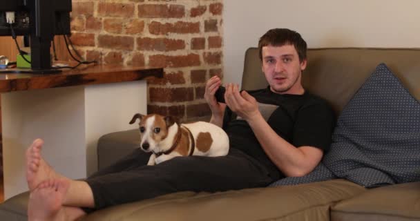 Мужчина смотрит на смартфон, сидящий на диване с собакой — стоковое видео
