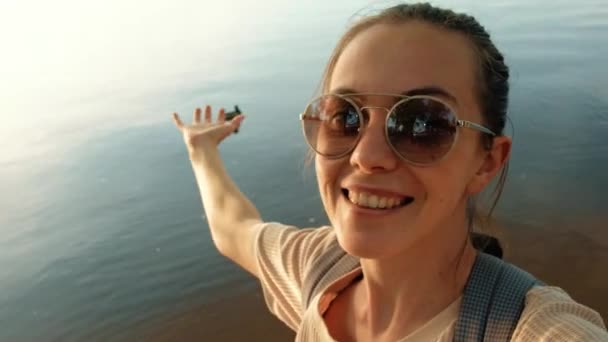 Щаслива молода жінка в сонцезахисних окулярах на березі проти спокійної води річкового шоку — стокове відео