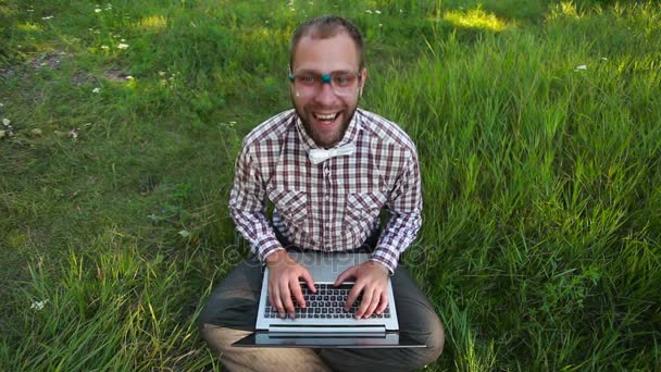 Junger Mann mit Brille sitzt auf dem Gras und tippt — Stockvideo