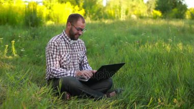 Çimenlikte oturan ve üzerinde lapto yazarak gözlüklü genç adam