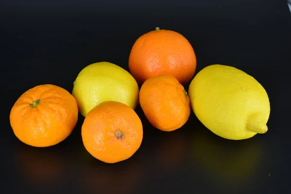 Fresh bright fruits, two yellow lemons, three small orange mandarins, one large mineralola lies on a black plastic background. Delicious and sweet fruits, fruit vase, healthy food for every day.