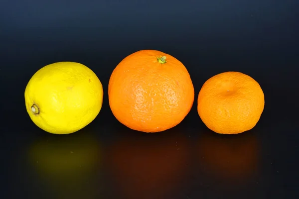 Fresh bright fruit, one yellow lemon, one small orange mandarin, one large mineralola lies on a black plastic background. Delicious and sweet fruits, fruit vase, healthy food for every day.