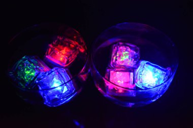 Siyah bir arka planda, farklı renkte parlak buz küpleri ve lezzetli bir içecek bulunan büyük bardaklar bulunur..