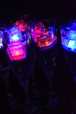 Farklı renklerde parlayan buz küpleri olan uzun burkulmuş bacaklarda beş cam kadeh. Aydınlık elementlere sahip eğlenceli ve parlak içecekler siyah bir arka plan, parti ve rahatlama üzerinde yer alır..