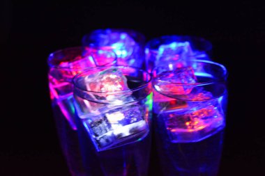 Farklı renklerde parlayan buz küpleri olan uzun burkulmuş bacaklarda beş cam kadeh. Aydınlık elementlere sahip eğlenceli ve parlak içecekler siyah bir arka plan, parti ve rahatlama üzerinde yer alır..